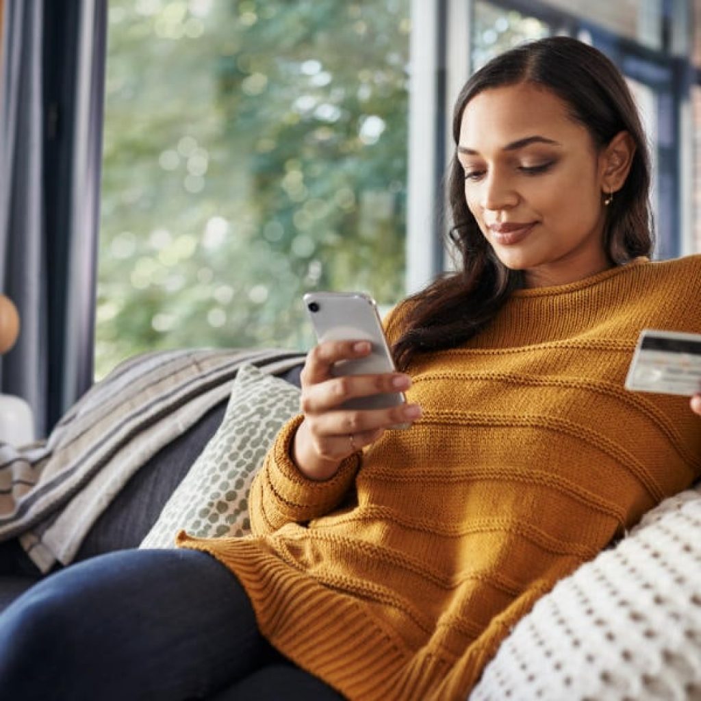 Shot of a beautiful young woman using her cellphone and credit card while relaxing on a couch at home